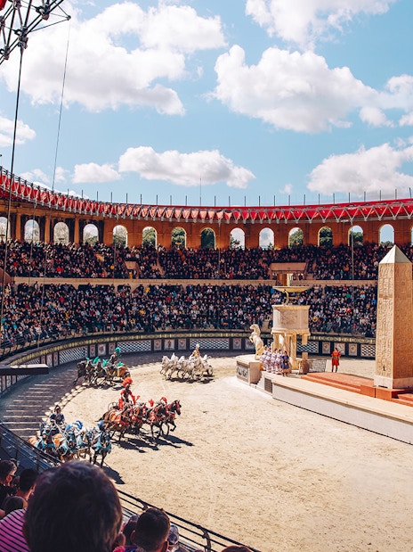 Chariot race in Puy du Fou arena with large audience and ancient Roman architecture.