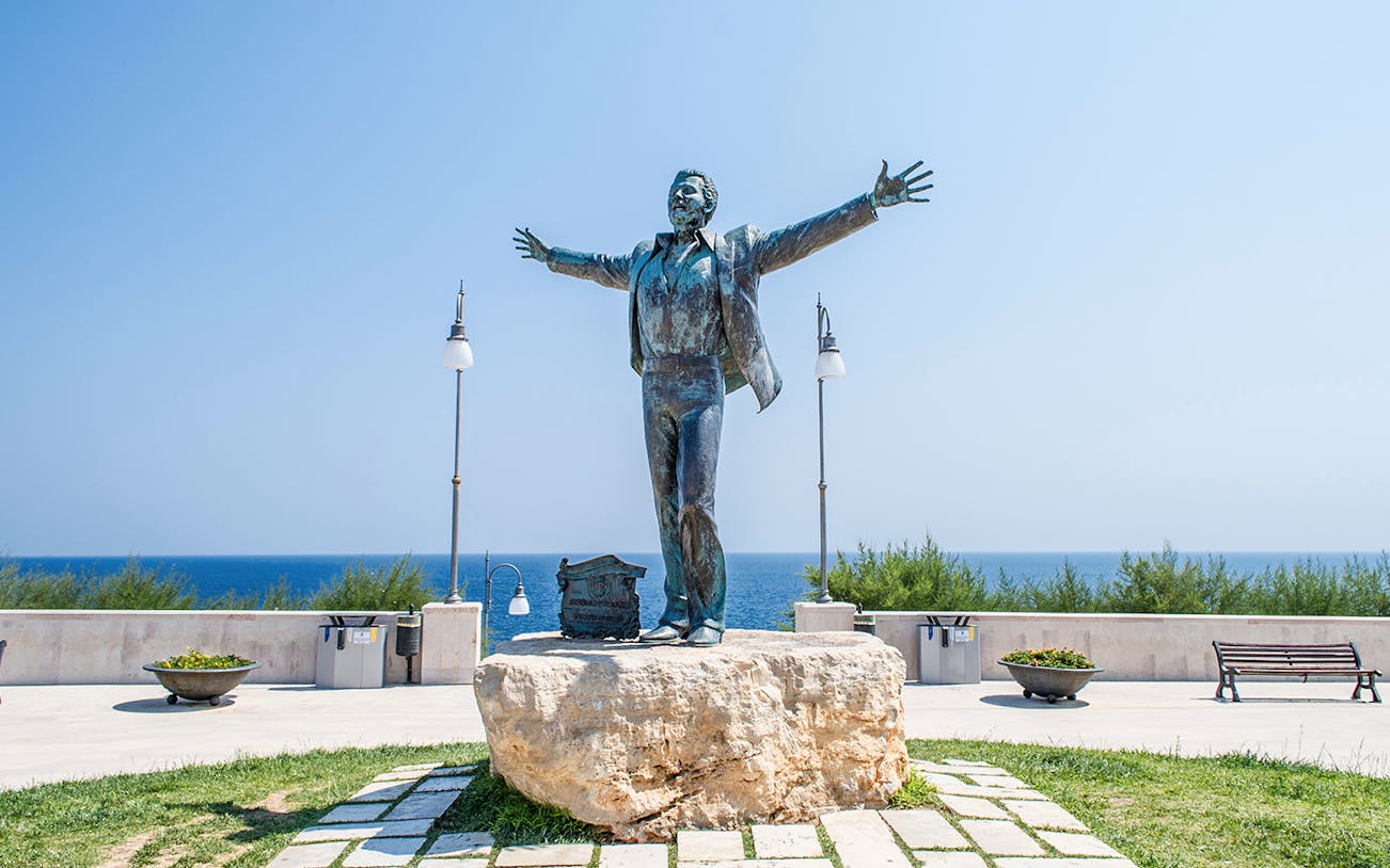 Statue of a man with open arms on a stone pedestal by the sea in Polignano a Mare.