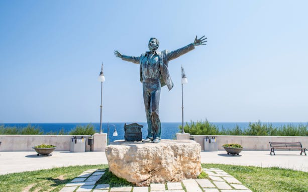 Statue of a man with open arms on a stone pedestal by the sea in Polignano a Mare.