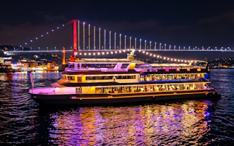 Bosphorus dinner cruise ship illuminated at night with Istanbul bridge in the background.
