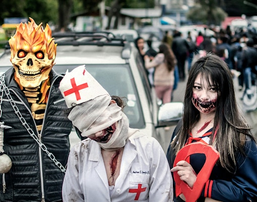 Costumed participants at a Halloween parade with a fiery skull, bandaged nurse, and zombie makeup.