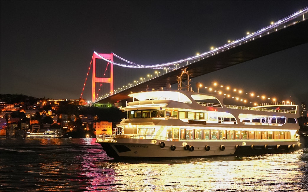 Bosphorus dinner cruise ship passing under illuminated Bosphorus Bridge at night.