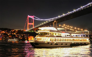 Bosphorus dinner cruise ship passing under illuminated Bosphorus Bridge at night.