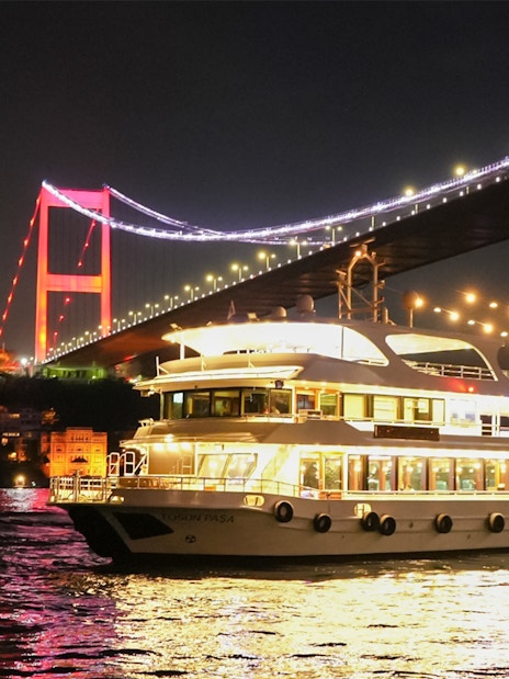 Bosphorus dinner cruise ship passing under illuminated Bosphorus Bridge at night.