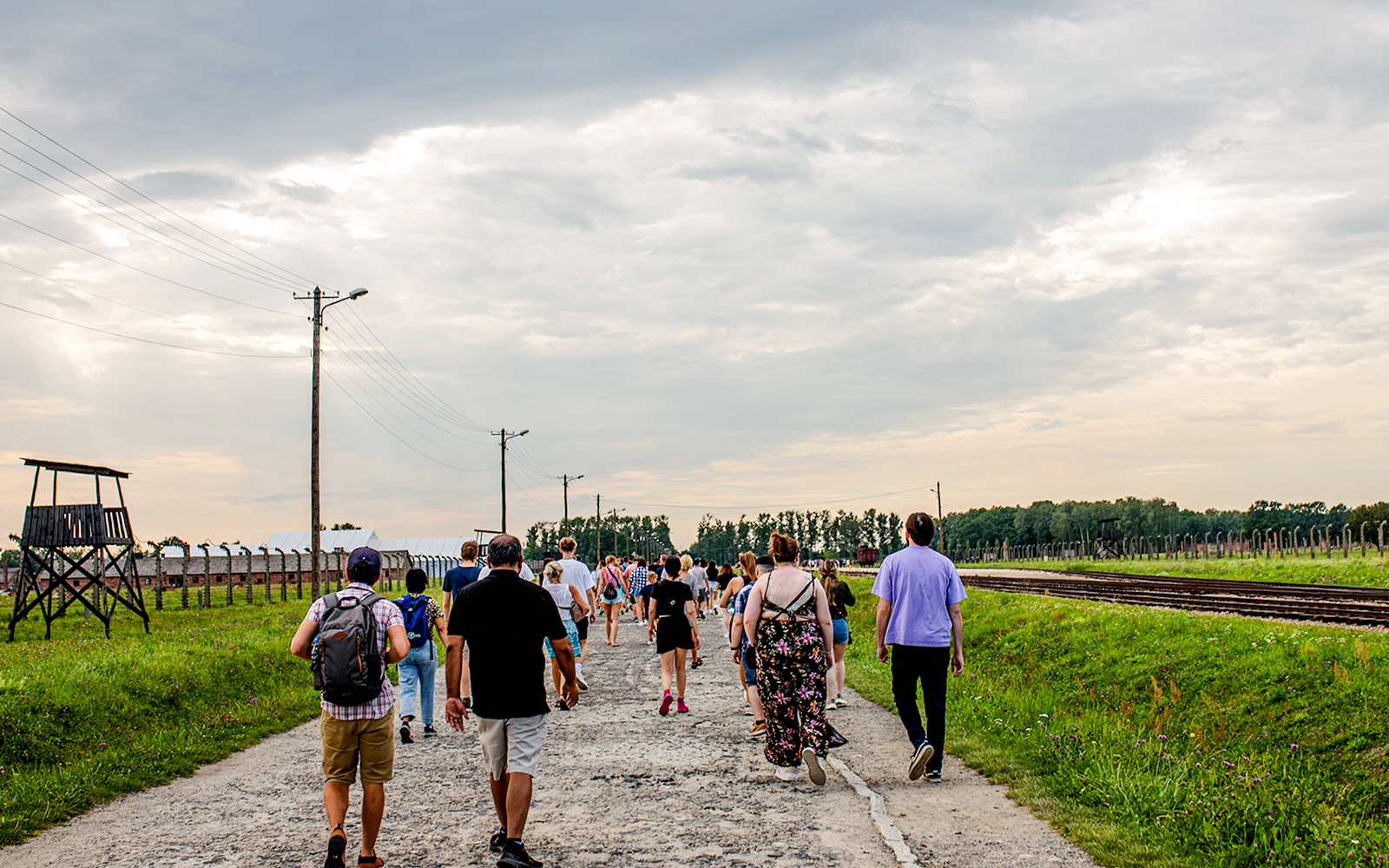 Visitors walking on a guided tour at Auschwitz-Birkenau, with watchtower and railway tracks visible.