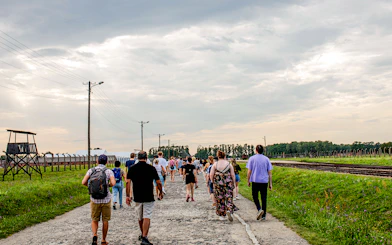 Visitors walking on a guided tour at Auschwitz-Birkenau, with watchtower and railway tracks visible.