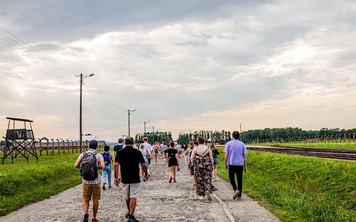 Visitors walking on a guided tour at Auschwitz-Birkenau, with watchtower and railway tracks visible.