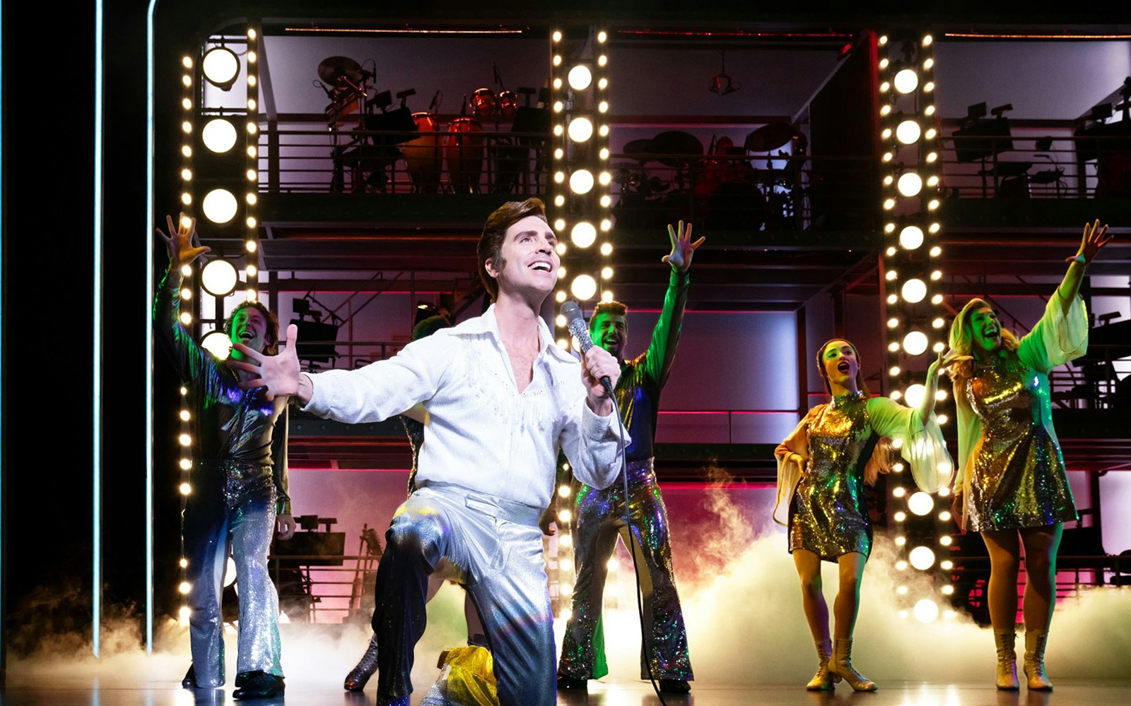 Performer singing on stage in "A Beautiful Noise: The Neil Diamond Musical" with vibrant lights and dancers.