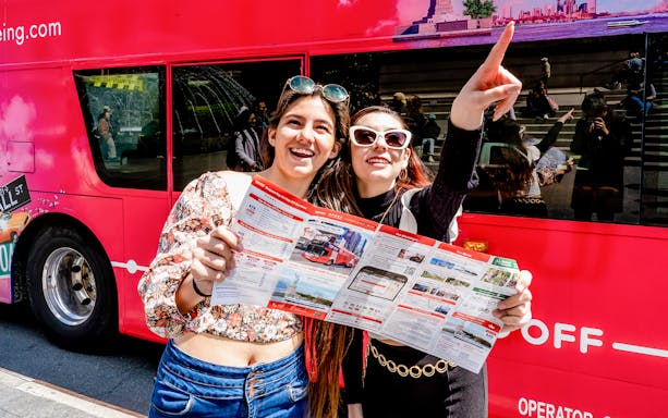 Tourists with map in front of red hop-on hop-off bus, pointing at city sights.