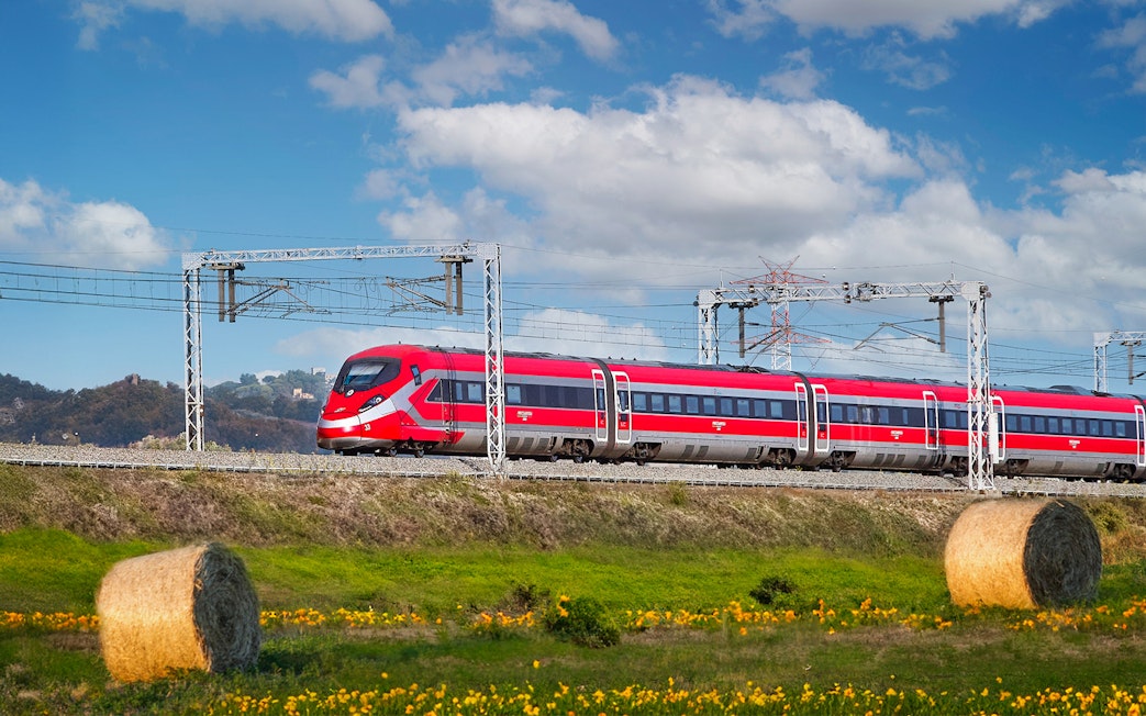 Trenitalia train traveling through Italian countryside with hay bales and hills.