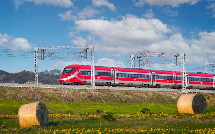 Trenitalia train traveling through Italian countryside with hay bales and hills.