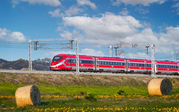 Trenitalia train traveling through Italian countryside with hay bales and hills.