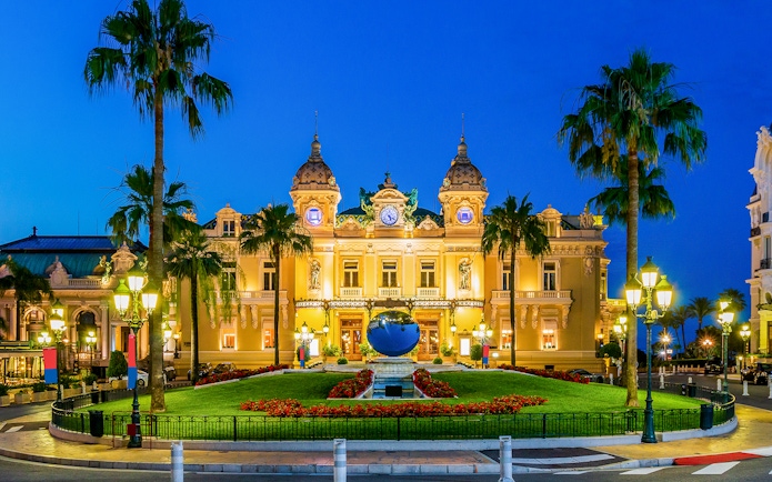 Monaco Casino illuminated at night with palm trees and gardens in the foreground.