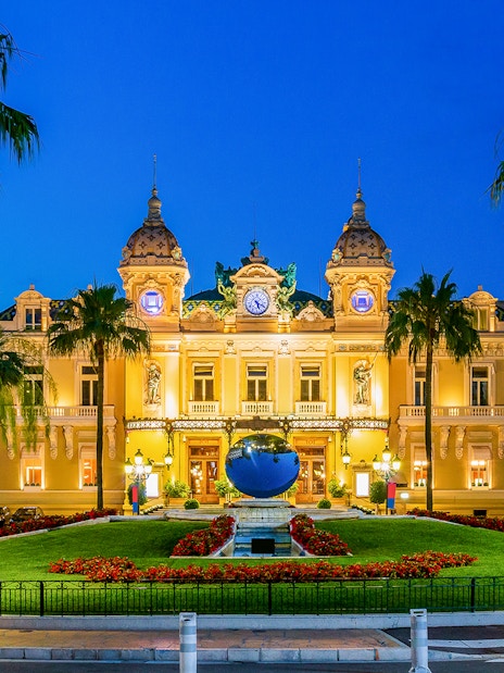 Monaco Casino illuminated at night with palm trees and gardens in the foreground.