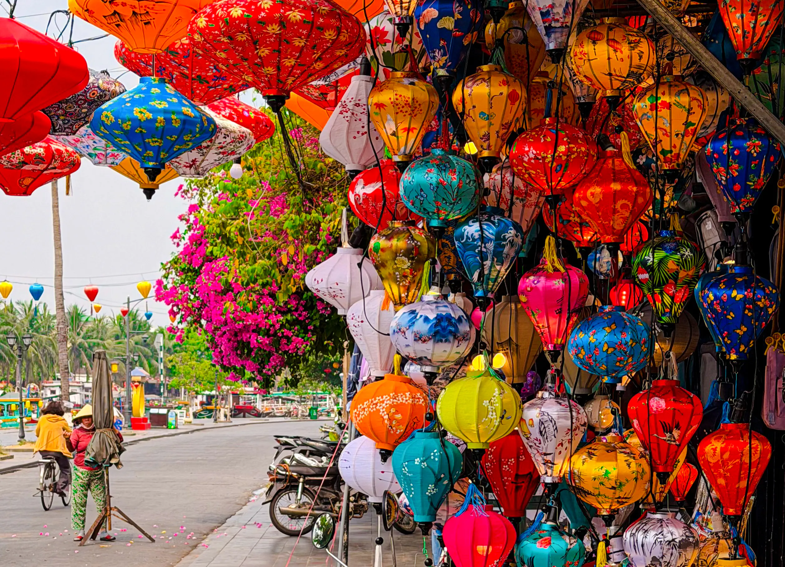 Colorful lanterns hanging in Hoi An street, Da Nang, with vibrant flowers and a cyclist nearby.