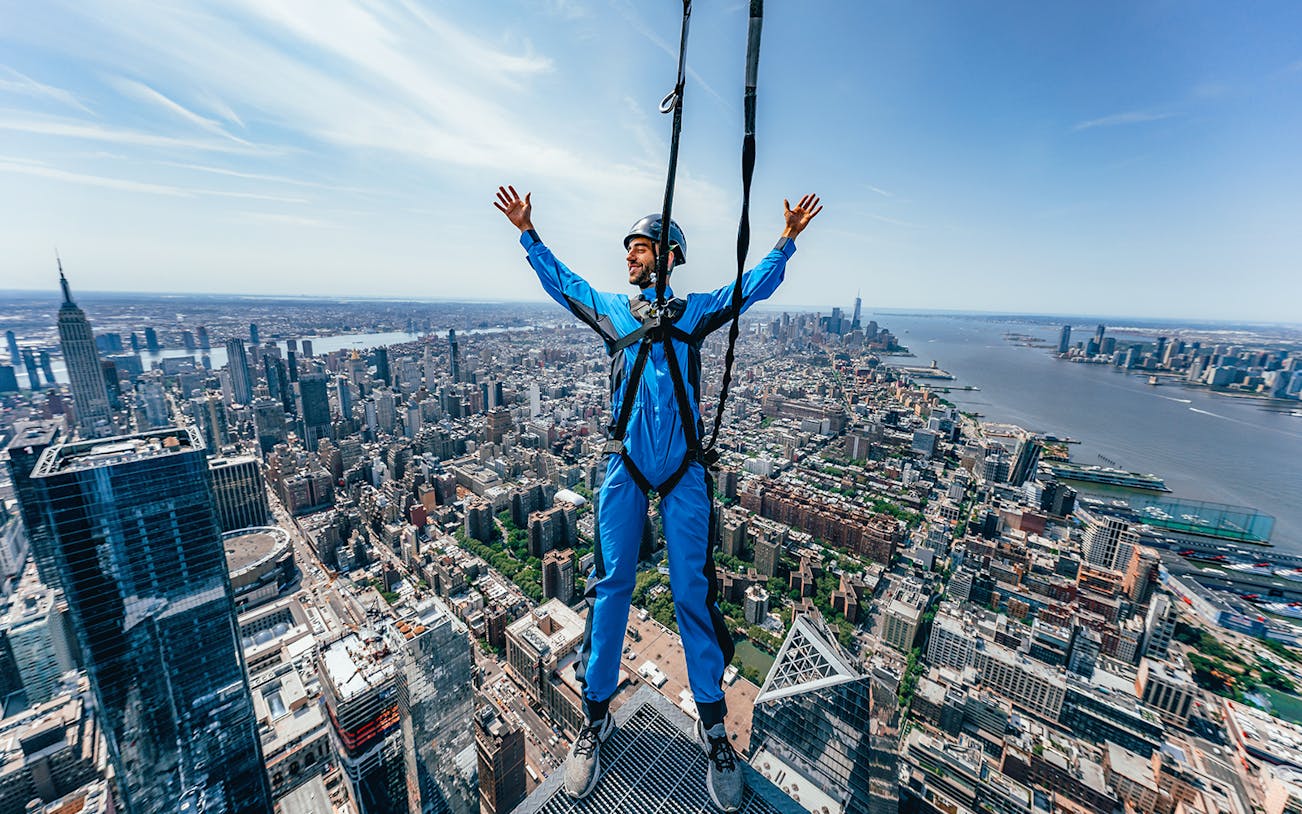 Person harnessed on skyscraper edge overlooking New York City skyline.