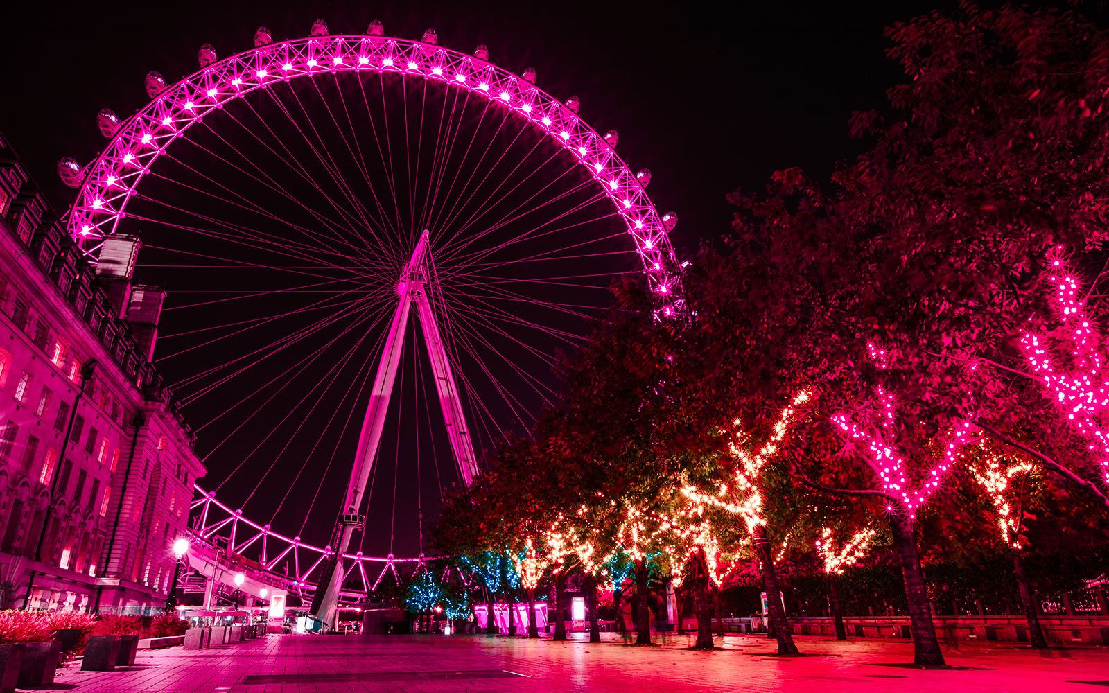 London Eye illuminated at night with vibrant pink lights, trees adorned with festive lights.