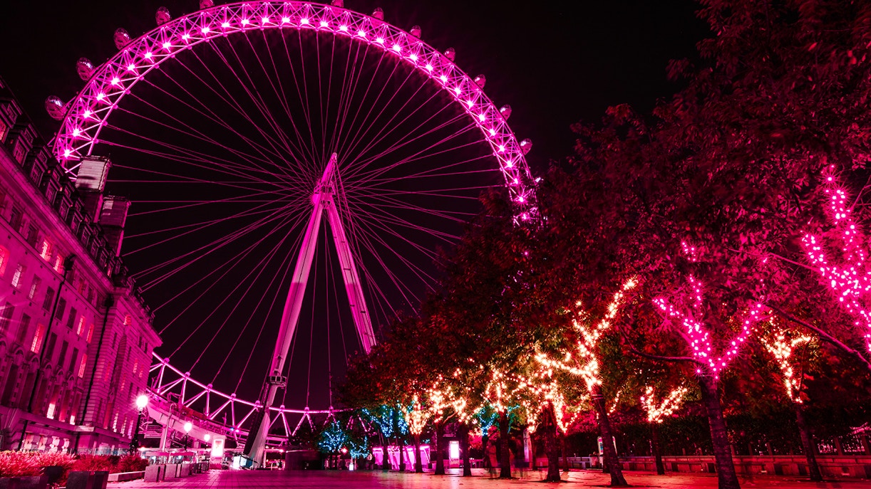London Eye illuminated at night with vibrant pink lights, trees adorned with festive lights.