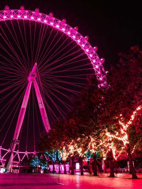 London Eye illuminated at night with vibrant pink lights, trees adorned with festive lights.