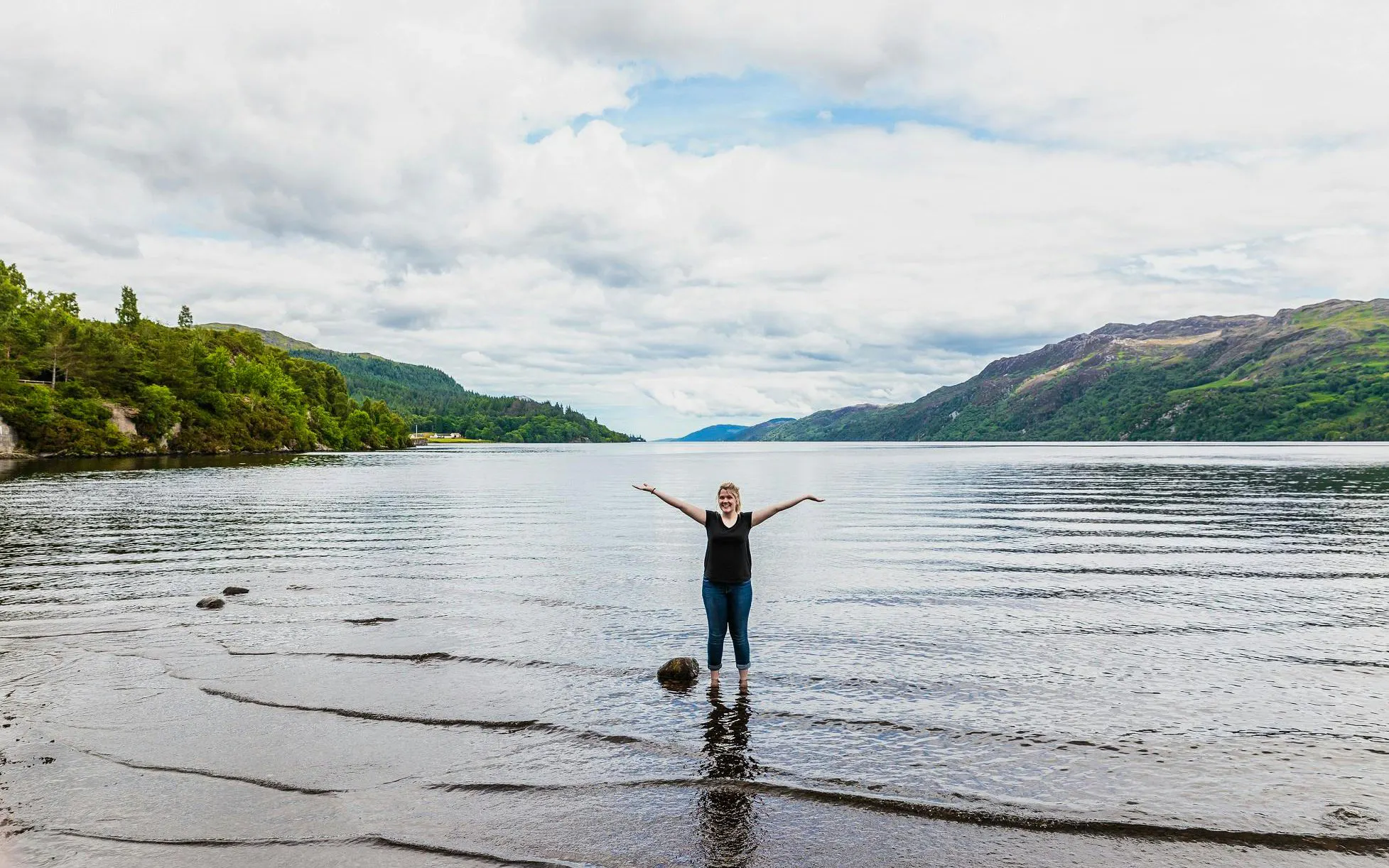Person standing in Loch Ness with arms raised, surrounded by hills and water.