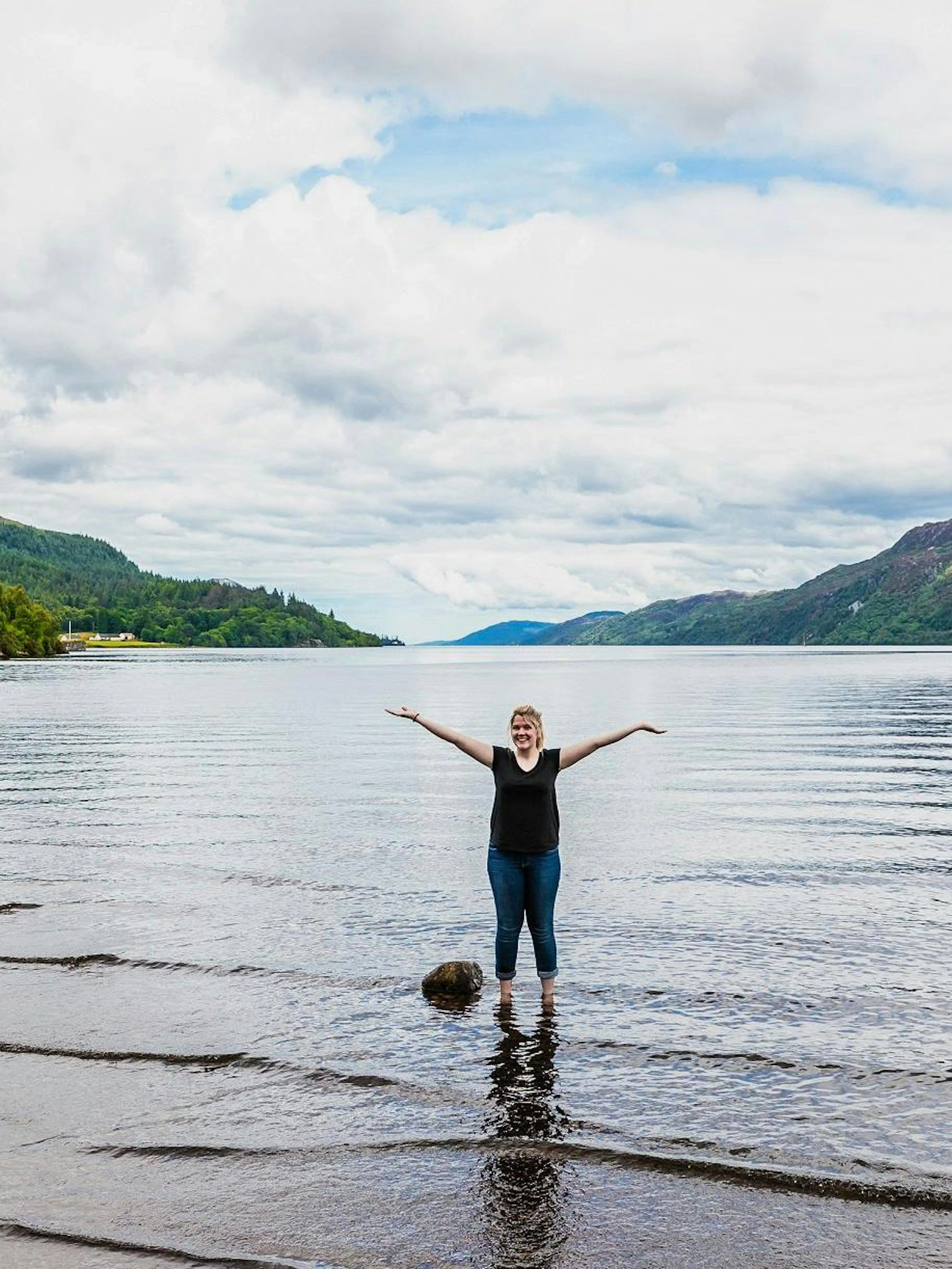 Person standing in Loch Ness with arms raised, surrounded by hills and water.