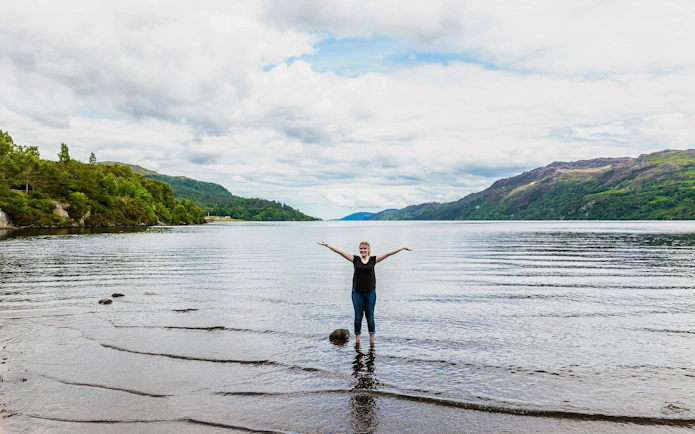 Person standing in Loch Ness with arms raised, surrounded by hills and water.