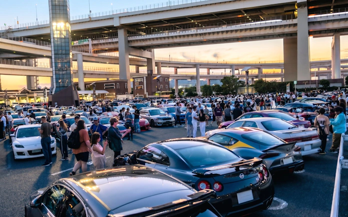 Car enthusiasts gather at Daikoku PA in Tokyo, showcasing diverse vehicles under highway overpasses.