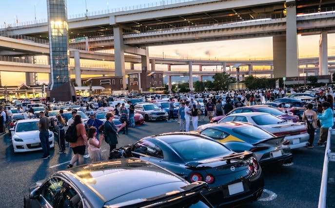 Car enthusiasts gather at Daikoku PA in Tokyo, showcasing diverse vehicles under highway overpasses.