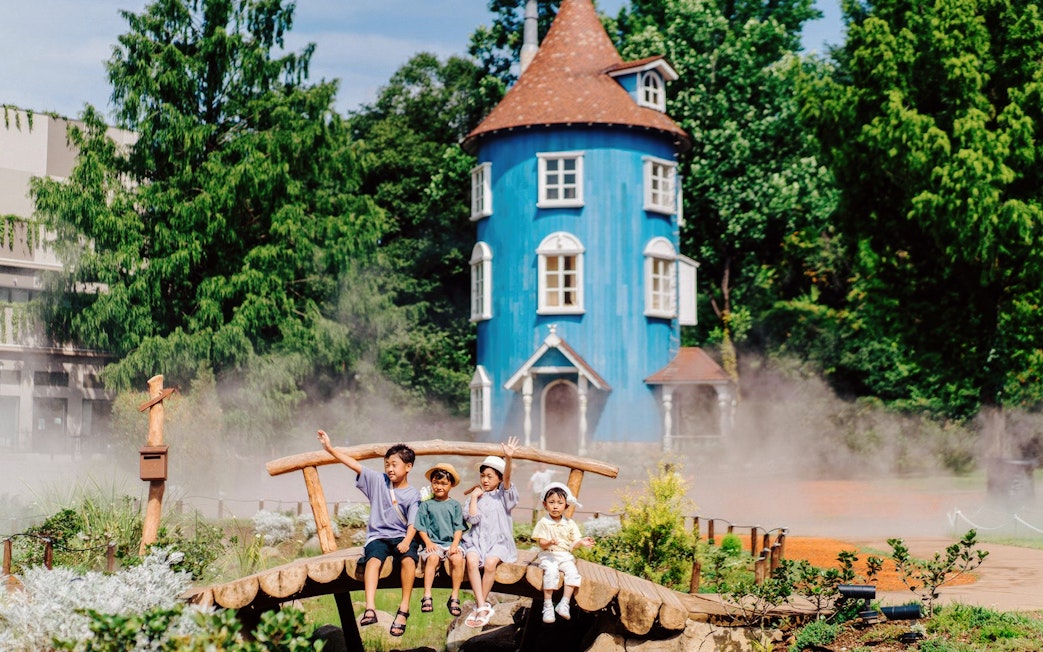 Children sitting on a wooden bridge in front of the blue Moominhouse at Moominvalley Park.