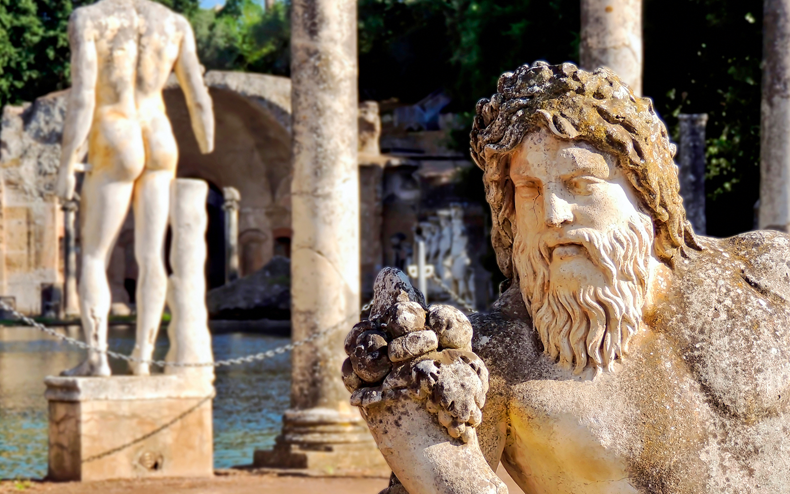 Statues at Villa Adriana, Tivoli, with ancient columns and water in the background.