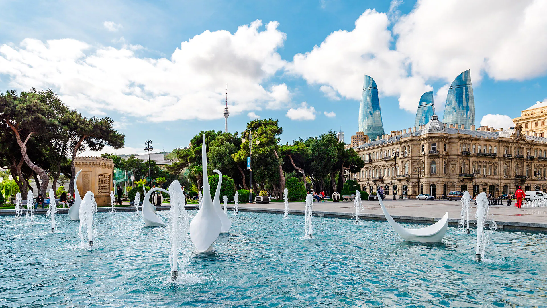 Fountain with sculptures in front of historic building and Flame Towers in Baku.