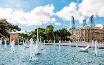 Fountain with sculptures in front of historic building and Flame Towers in Baku.