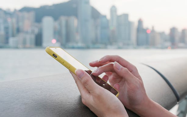 Hands using smartphone with Hong Kong skyline in background, highlighting travel connectivity.