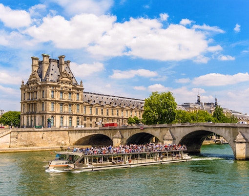 Louvre Museum entrance with visitors and Seine River cruise boat in Paris.