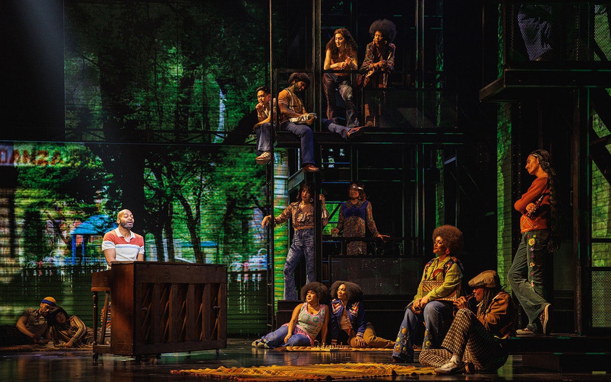 Stage performance in Hell's Kitchen with actors in 1970s attire, featuring a pianist and ensemble cast.