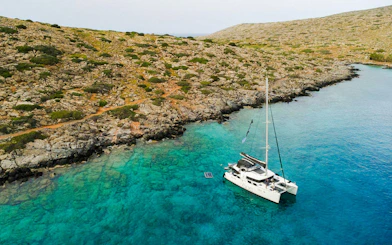 Catamaran anchored in clear waters near rocky Heraklion coastline.