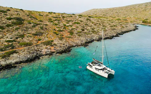 Catamaran anchored in clear waters near rocky Heraklion coastline.
