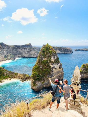 Young family admiring Thousand Islands Viewpoint, Nusa Penida, Bali, with ocean and cliffs in background.