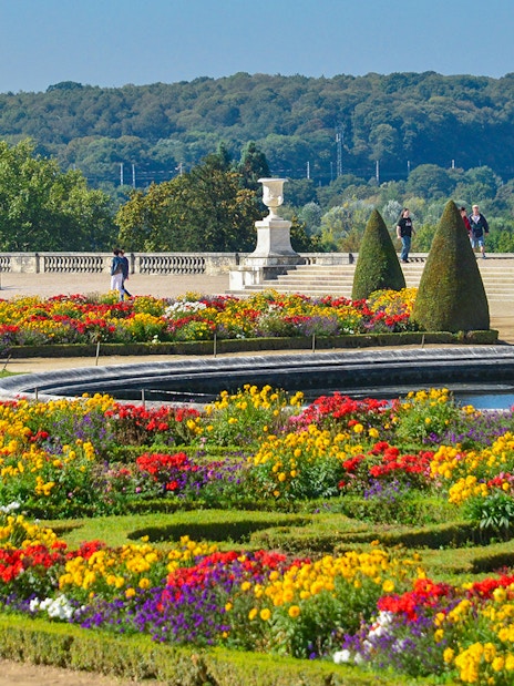 Tourists walking in Versailles Palace garden with vibrant flower beds.