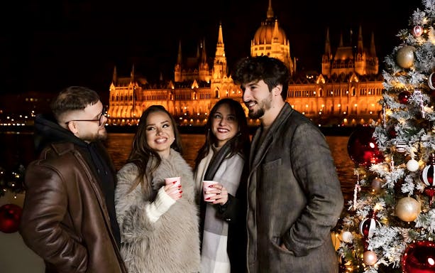 People enjoying a Christmas Market Cruise in Budapest with the Parliament building illuminated at night.