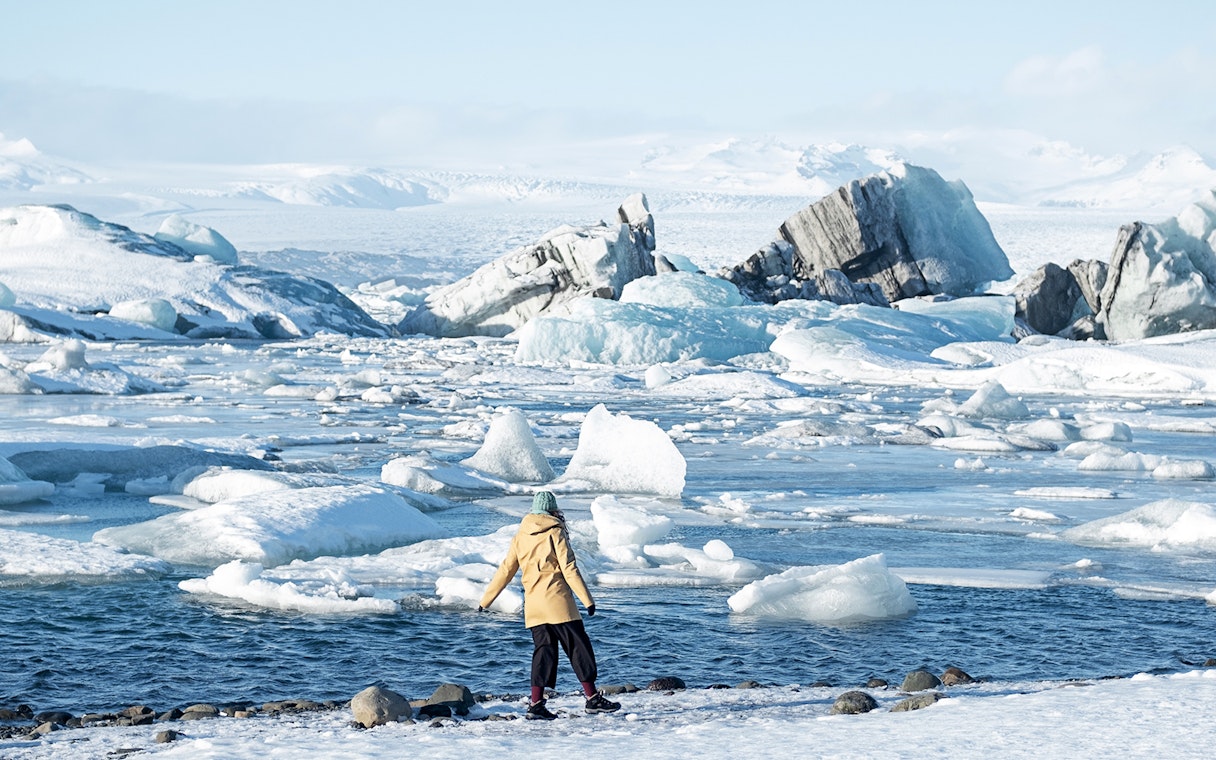 Person standing by icy waters at Jökulsárlón Glacier Lagoon, Iceland, with icebergs in the background.