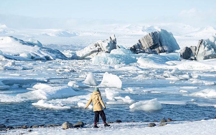 Person standing by icy waters at Jökulsárlón Glacier Lagoon, Iceland, with icebergs in the background.