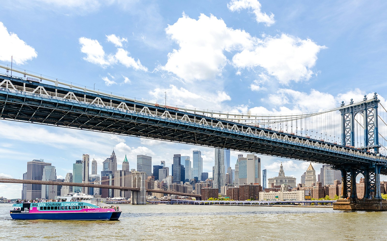 Cruise boat under Manhattan Bridge with New York City skyline in the background.