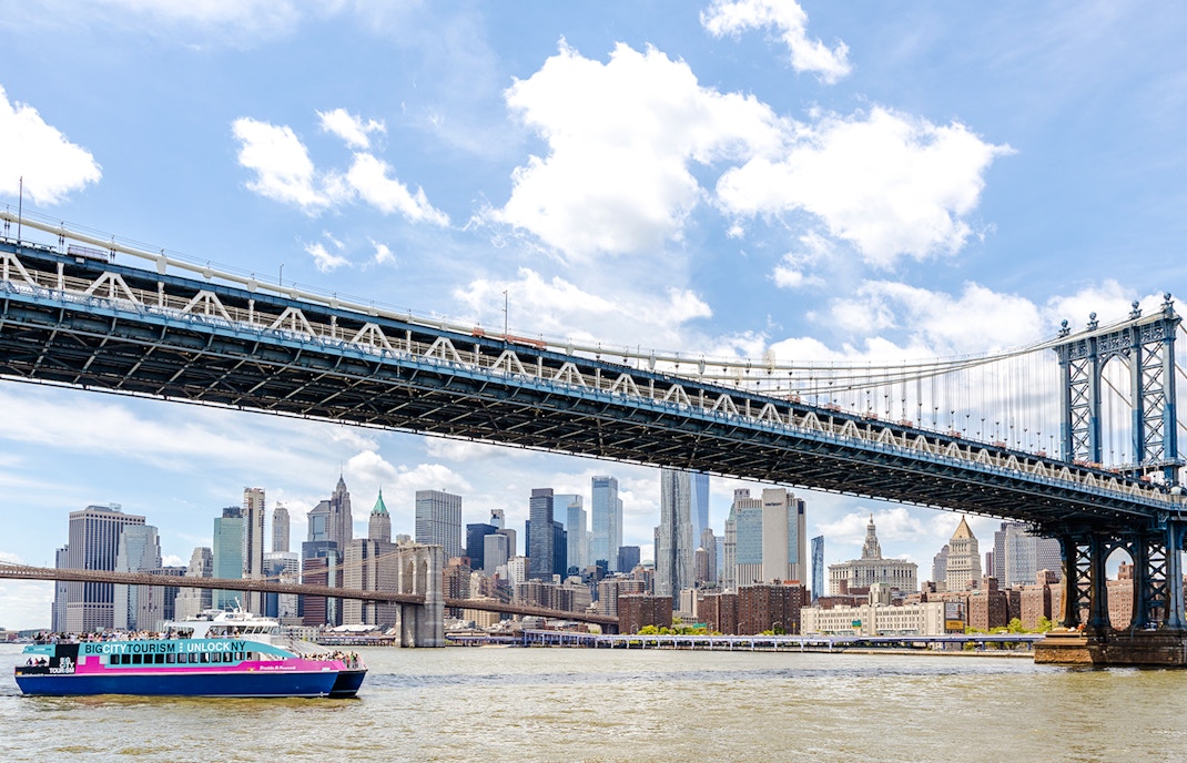 Cruise boat under Manhattan Bridge with New York City skyline in the background.