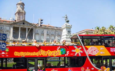 People on a hop-on hop-off bus in Porto, passing by the Palácio da Bolsa and a statue.
