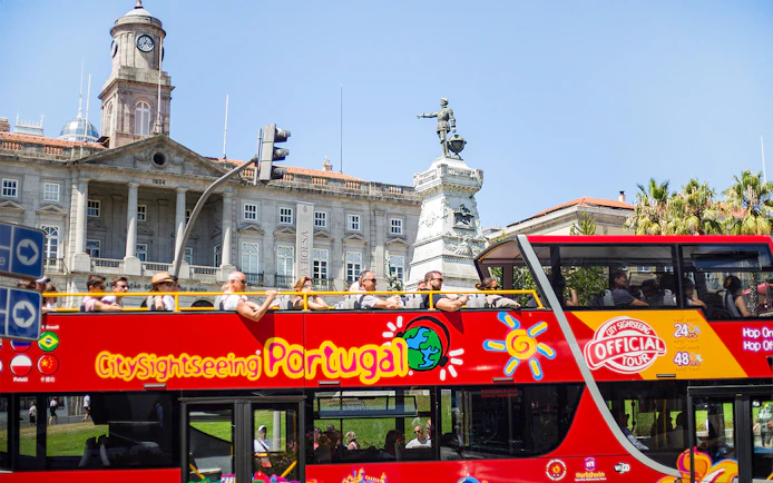 People on a hop-on hop-off bus in Porto, passing by the Palácio da Bolsa and a statue.