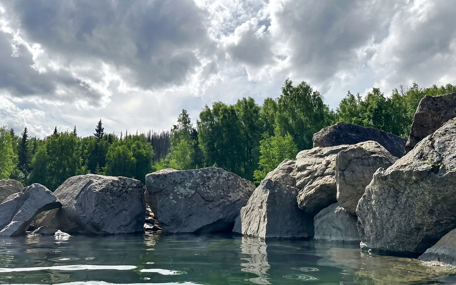 Rocky shoreline and lush trees at Chena Hot Springs, Alaska, under a cloudy sky.