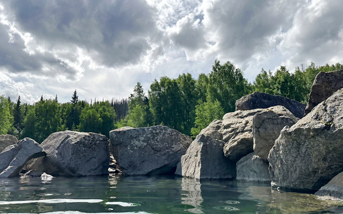 Rocky shoreline and lush trees at Chena Hot Springs, Alaska, under a cloudy sky.