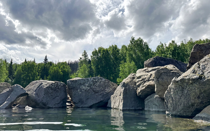 Rocky shoreline and lush trees at Chena Hot Springs, Alaska, under a cloudy sky.