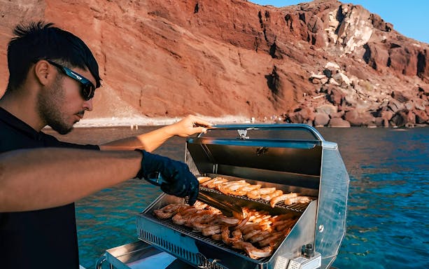 Grilling seafood on a boat near Santorini's red cliffs.